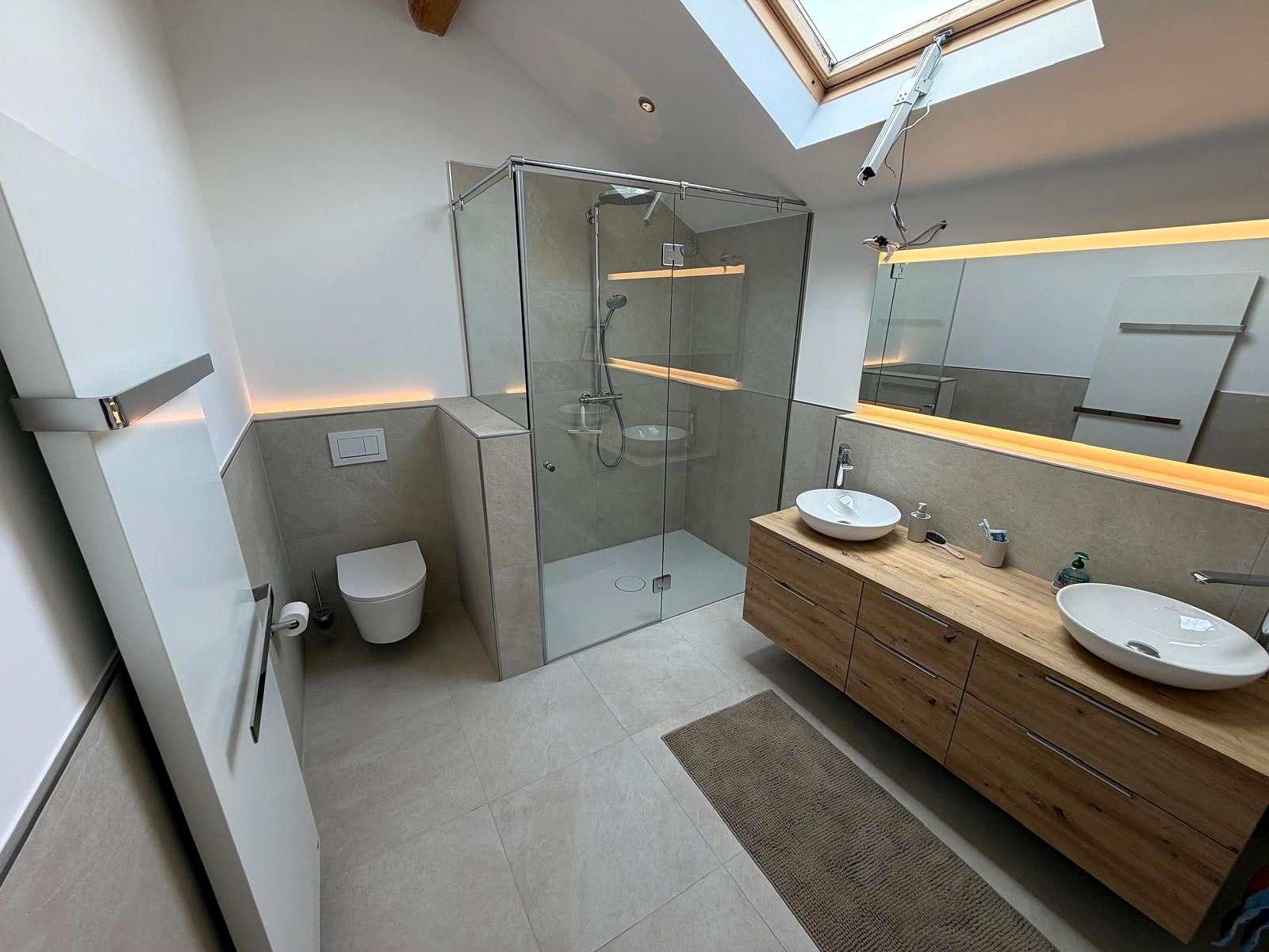 Dachgeschossbad mit Glasdusche, beleuchteter Nische, Doppelwaschtisch aus Holz und Fliesen in Betonoptik