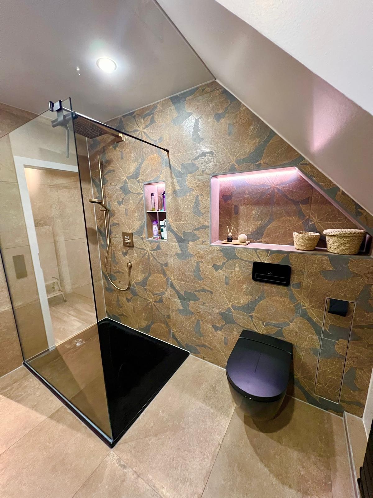 Dachgeschossbad nach der Sanierung: großformatige Fliesen mit Blattmotiv, Glasdusche mit schwarzer Duschwanne, beleuchtete Wandnische und schwarzes Wand-WC